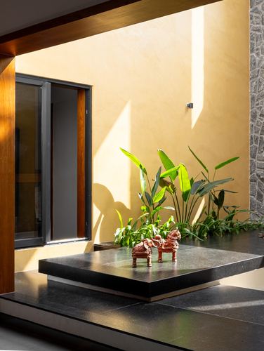 Sunlit courtyard corner with mustard wall, banana plant, and dhokra figurines on a black stone platform