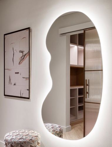 Organic backlit mirror on a beige wall reflecting a walk-in closet with fluted glass door and wood shelving