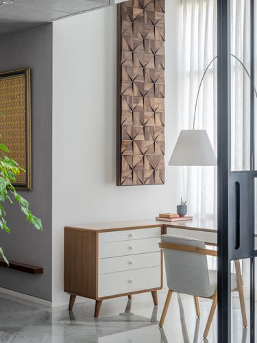 Study nook with wood-paneled desk, cane chair, chest of drawers, and geometric wood wall art beside curtained window