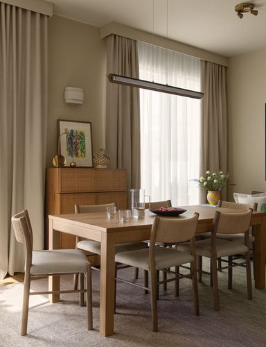 Wood dining table with cane chairs beneath a linear pendant light near curtained window