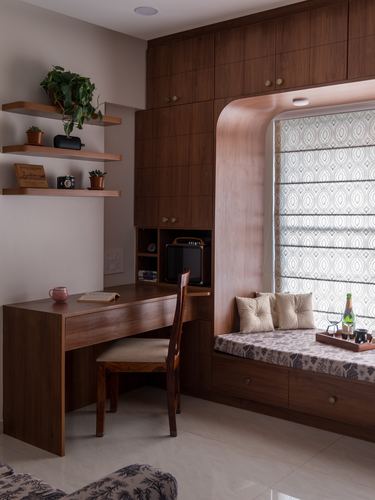 Wooden study desk beside a curved archway window seat with patterned cushions and blinds