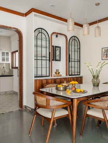 Dining room with cane teak chairs, marble-top table, marigolds, and arched glass cabinet doors under pendant lights