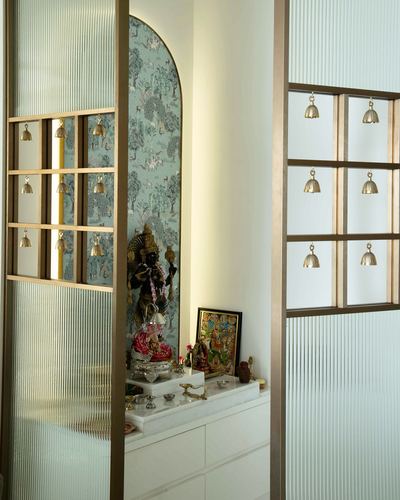 Pooja niche with deity idol framed by arched patterned wall, seen through reeded glass and brass grille doors hung with bells