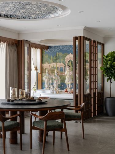 Round dining table with teak chairs opens through a wooden screen to a mural-backed pooja alcove
