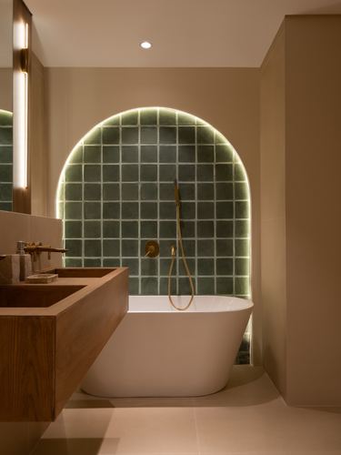 Freestanding tub beneath backlit arched green tile niche with brass fixtures in beige bathroom