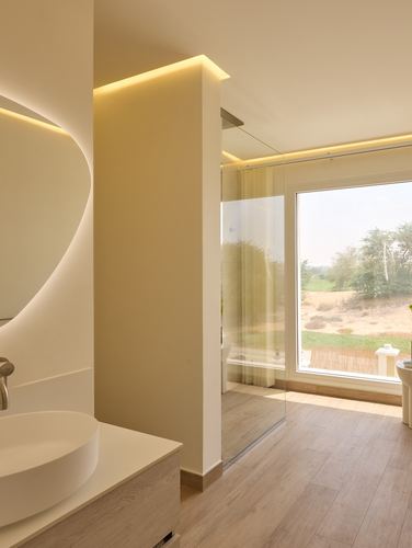 Modern bathroom with curved backlit mirror, wood floor, glass shower, and window view to landscape.