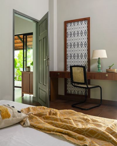 Bedroom corner with cane chair at wood desk, ikat panel, open green door to greenery beyond