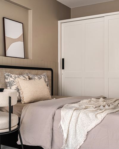 Bedroom corner with cane headboard, layered cushions, and white sliding wardrobe against beige walls.