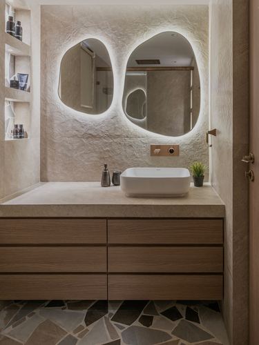 Bathroom vanity with two organic backlit mirrors, stone counter, and wood drawers over pebble floor