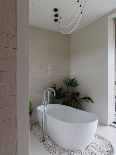 Oval freestanding tub on pebbles beneath sculptural pendant lights and tiled wall