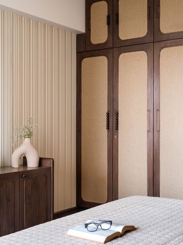 Wardrobe with fabric-inset wood doors beside fluted wall panel and bed in warm-toned bedroom