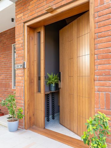 Wooden pivot front door open in a brick facade with a fern-topped console visible inside