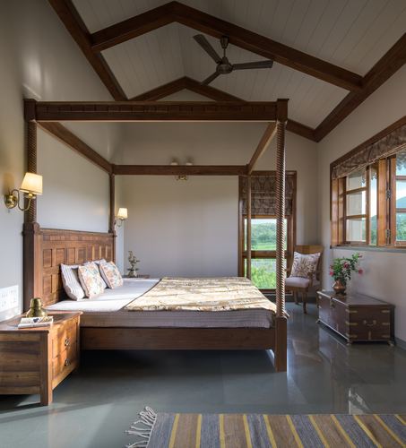 Teak four-poster bed under exposed wooden roof trusses in a farmhouse bedroom with stone flooring
