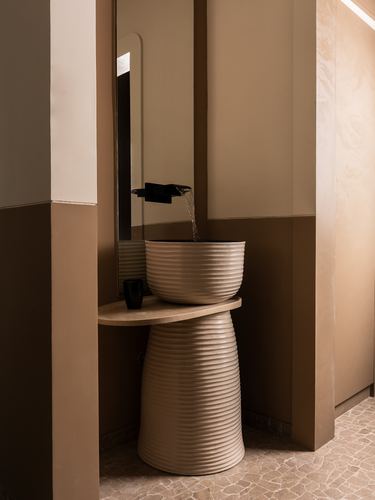 Ribbed ceramic pedestal basin with black faucet against mirror in warm brown-toned bathroom niche