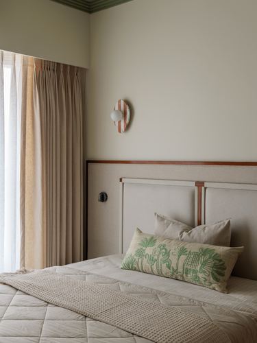 Bedroom corner with upholstered headboard, green embroidered pillow, pleated curtains and a striped wall sconce