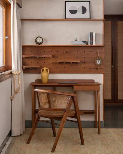 Wooden study desk with cane chair beneath a wall-mounted pegboard shelf and framed print