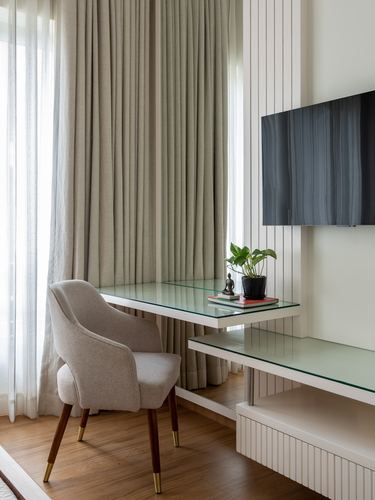 Beige fluted-panel study nook with glass desk, mounted tv, and cane-less upholstered chair