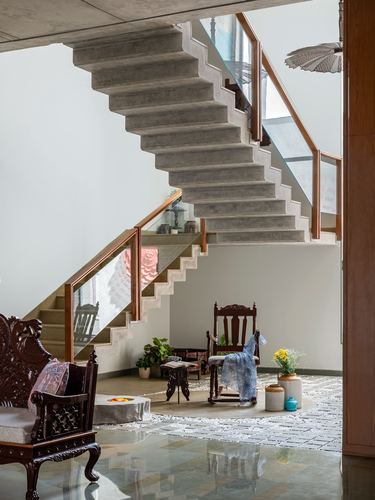 Cantilevered concrete staircase above a seating nook with carved wooden chairs and stone flooring