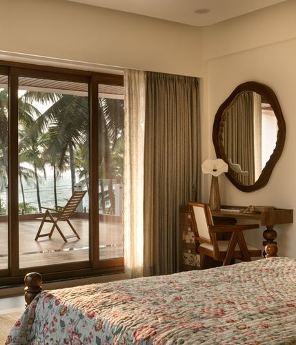 Bedroom with floral quilt, wooden desk and round mirror beside glass door opening to sea-view balcony