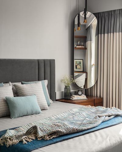 Grey upholstered bed with teal patterned throw beside wooden nightstand and tall oval mirror