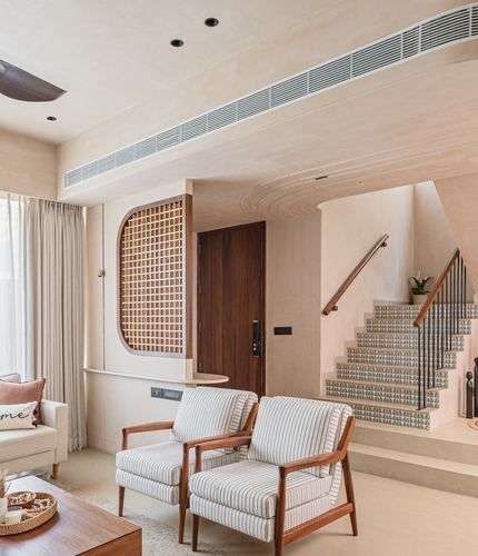 Living room with striped armchairs facing a curved lattice screen beside a tiled staircase and wooden door