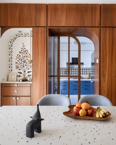 Dining table with fruit bowl in front of wood-paneled wall with arched niche and view into blue kitchen