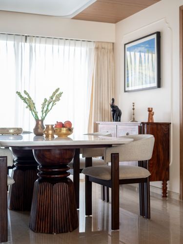 Dining table with carved wood pedestal base, marble top, and upholstered chairs beside a sunlit window