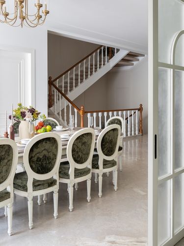 Dining table with oval-back green velvet chairs beside a wooden staircase with white balusters