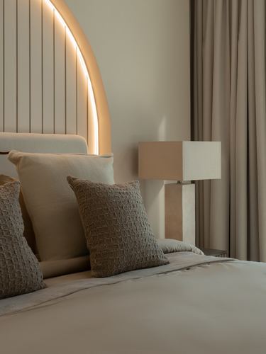 Bedside vignette with arched upholstered headboard, backlit cove lighting, textured cushions, and stone table lamp