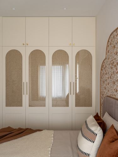 Cream wardrobe with arched glass panel doors beside a bed with terracotta cushions and patterned headboard wall