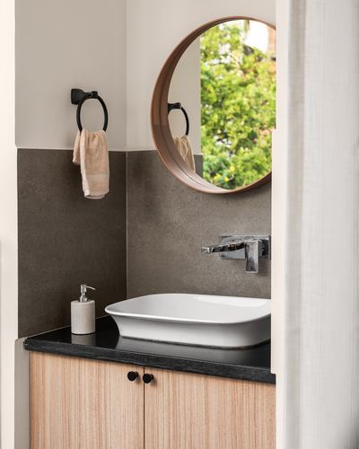 Bathroom corner with round wooden mirror, white vessel sink, black stone counter and wood cabinetry.