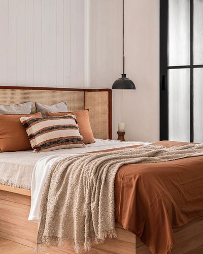Bed with cane headboard, terracotta linens and bouclé throw beside black pendant light and reeded glass door