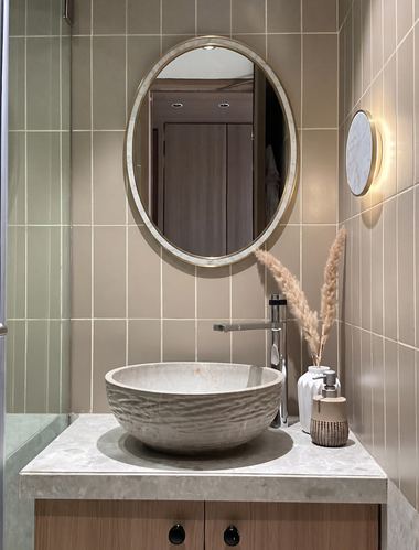 Oval mirror above a textured stone vessel sink on a wood vanity in a tiled bathroom