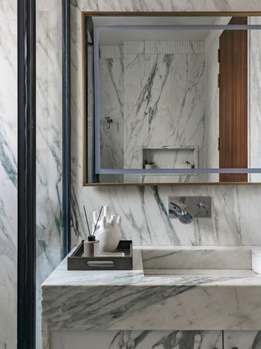Marble bathroom vanity with backlit mirror reflecting shower niche and wood door