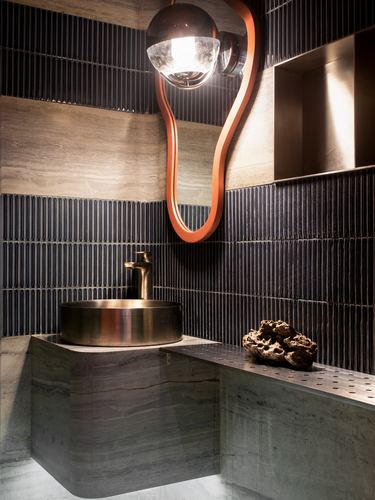 Bathroom with ribbed dark tile walls, brass vessel sink on stone counter, sculptural orange mirror and pendant light