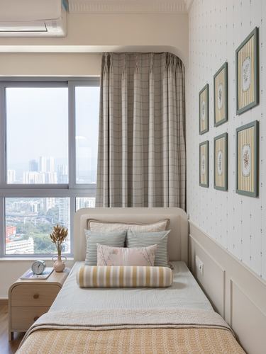 Single bed with layered pastel cushions beneath a framed gallery wall, city view through window