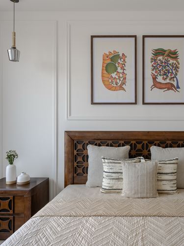 Bed with carved wood headboard, layered cushions, and framed folk art above on paneled wall