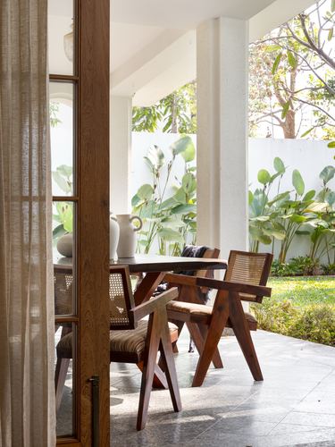Covered veranda with cane-and-wood chairs at a table, framed by curtain and garden beyond