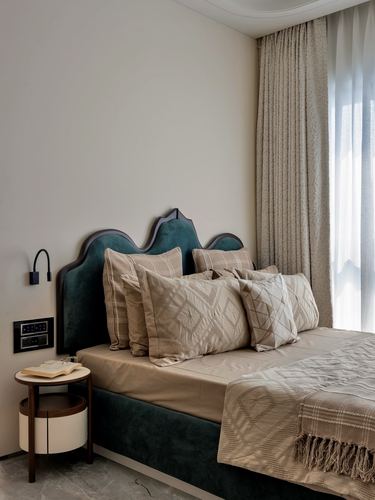 Bedroom corner with curved teal velvet headboard, layered beige pillows, and round bedside table
