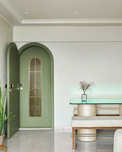 Sage green arched entrance door with brass grille inset beside fluted white wall and glass dining table