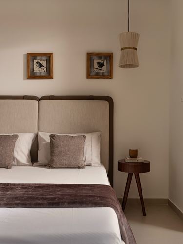 Bedroom with beige leather-trimmed headboard, framed bird prints, woven pendant light and round wooden side table