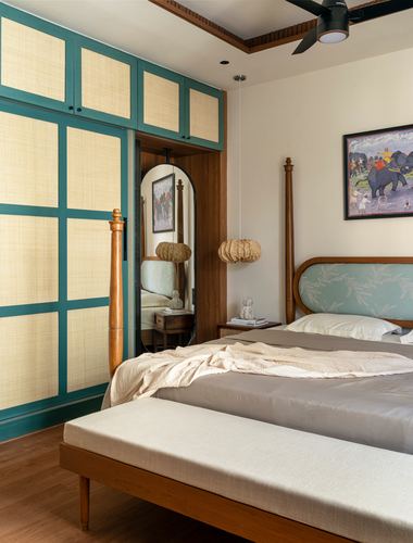 Bedroom with cane-panelled teal wardrobe, four-poster wooden bed, arched mirrored doorway, elephant painting on wall.