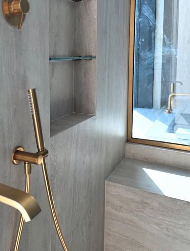 Close-up of brushed brass shower fixtures against a stone-clad wall with a recessed niche and window