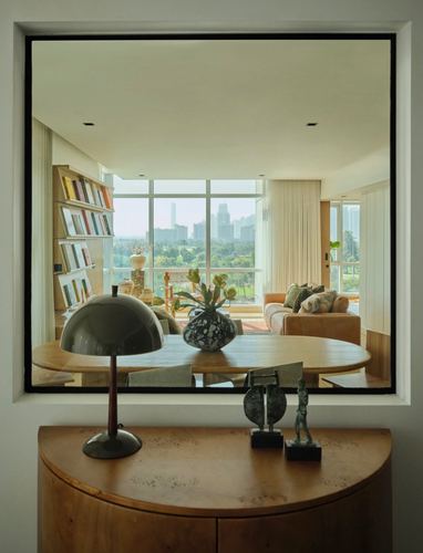 Wooden console with sculptural lamp beneath a black-framed opening revealing a sunlit living room and city skyline.