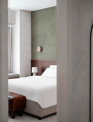 Bedroom glimpsed through a doorway, sage plaster wall, fluted headboard panel, white bedding, leather ottoman