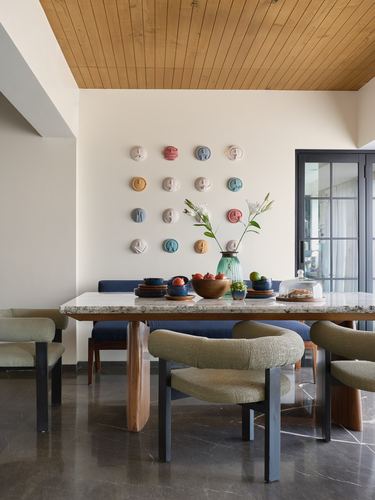 Dining room with marble table, olive chairs, navy banquette, wood ceiling, and ceramic face discs on wall