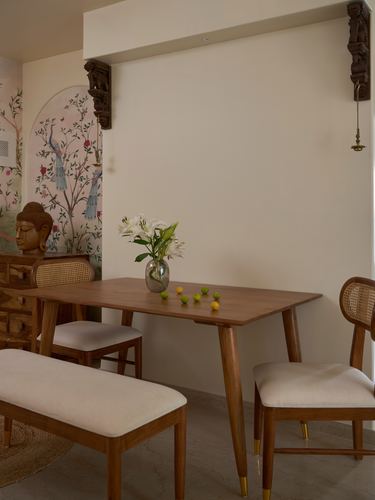 Teak dining table with cane chairs, bench, flowers, and carved wooden corbels against a cream wall