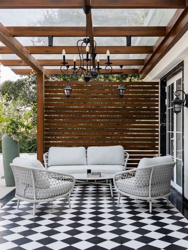Covered patio with checkerboard floor, wicker chairs, and wrought-iron chandelier under glass pergola