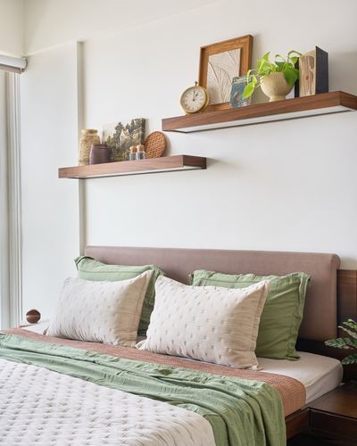 Bedroom with upholstered headboard, green and cream linens, and two wooden shelves styled with books and plants