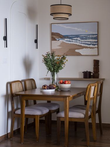 Wooden dining table with cane-back chairs beneath a framed seascape painting and floating shelf